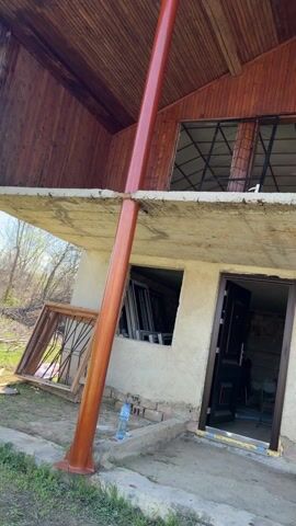 The video shows a two-story building under construction. The top floor has wood paneling and window openings. The ground floor is concrete with a doorway and window openings that are not yet completed. There are copper-colored support pillars. To the side of the building is a trench dug in a field. Two bottles of water are present. The weather appears sunny.