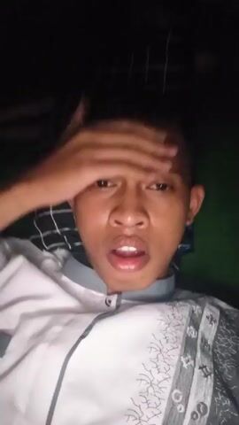 A young man is shown in a close-up shot, appearing to be lying down in a dark environment. He is wearing a white shirt with a gray pattern. The man is looking at the camera and makes a few gestures with his hands, including touching his hair and forming a peace sign. The lighting is dim, and the background is out of focus.