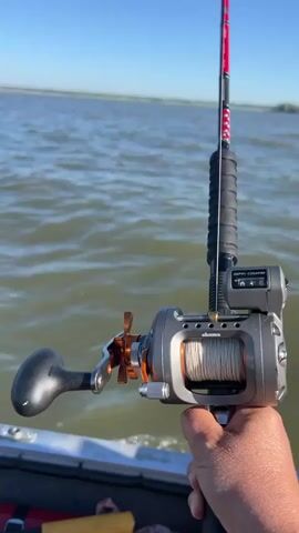 A first-person perspective video shows a person holding a fishing rod and reel while on a boat. The fishing reel is gray and orange with white fishing line. Attached to the rod above the reel is a digital depth counter. The boat is moving through water, creating a wake. The water is choppy and a clear blue sky is visible in the background with a distant horizon. A person's hand is visible holding the fishing rod.