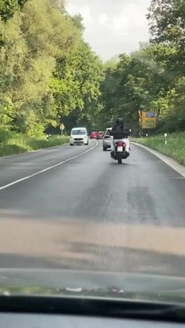 The video shows a view from inside a vehicle driving on a wet road with trees lining both sides. Several vehicles are visible on the road, including a white van, a white SUV, a silver car, and a black motorcycle with a rider. The motorcycle is in the foreground, and the rider is wearing a black helmet and jacket. Traffic appears to be moving slowly. A yellow road sign is visible in the distance on the right side of the road. The sky is overcast.