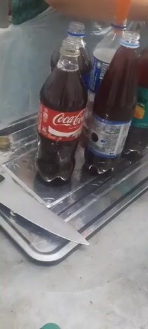 The video shows a close-up of several plastic bottles of soda and juice arranged on a metal tray. A knife is visible in the foreground, with its blade resting on the tray. The bottles are of various brands, including Coca-Cola, and some appear to be partially consumed. The lighting is dim, and the background is blurred, suggesting an indoor setting. The overall impression is one of preparation or a casual gathering.