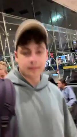 A young man, wearing a grey hoodie, a light colored baseball cap, and a backpack, is filming himself at what appears to be an airport. He is facing the camera, close up, with a slight smile. Other people are visible behind him, including a woman in an orange shirt and a man with bleached hair and a pink shirt. Some people are sitting in chairs along the wall to the right. The background shows large windows with a view of the night sky, and signage is visible, suggesting an airport concourse or waiting area. The camera is slightly shaky.
