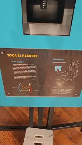 A person's hand reaches into a black rectangular display case containing a coiled metal spring. The hand moves towards the spring, appearing to be attempting to touch it. A sign below the display case has text in Spanish, and a diagram showing how light reflects.