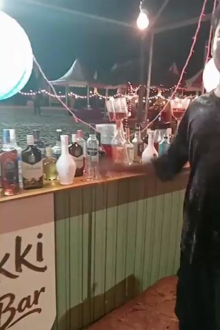 The video displays an outdoor bar setup at night. A light green, horizontally planked bar counter occupies the foreground, extending across the frame. On the counter, numerous bottles of various alcoholic spirits are arranged, alongside several small shot glasses. To the right of the bottles, three tall, clear beverage dispensers with spigots contain different colored liquids. A large white spherical lamp hangs above the right side of the bar, and red string lights are draped across the overhead structure. In the background, out of focus, multiple tents are visible, illuminated by string lights. The ground beneath and around the bar appears to be sand. The camera slowly pans from left to right, showcasing the entire bar area.