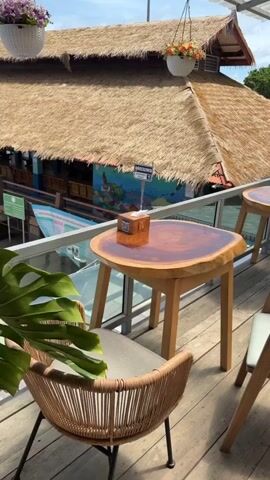 The video shows an outdoor seating area of a cafe or restaurant with a thatched roof structure visible in the background. The foreground features wooden tables and chairs. One table has a tissue dispenser. Hanging plants and potted plants are arranged around the seating area. The lighting suggests it is daytime with a clear sky. The overall atmosphere appears relaxed and natural.