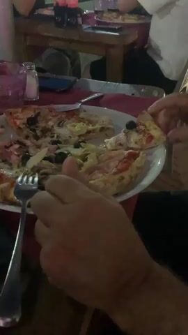 This is a close-up video of a person's hands and part of their face as they eat pizza at an outdoor restaurant. The person is holding a slice of pizza and bringing it to their mouth. In the background, other people are visible sitting at tables and eating. There are bottles of soda on a table in the background. The lighting suggests it is evening.
