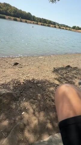 The video opens with a wide shot of a lake. In the foreground, there is a rocky, dirt shoreline with scattered leaves and shadows from trees. A person's legs, wearing white sneakers and black shorts, are visible on the right side of the frame. A straw hat is positioned on the left side of the frame, also in the foreground. The camera then pans to focus on a hand holding a paper cup filled with yellow, soft-serve ice cream, which appears to be mango flavored given the logo on the cup. The hand has painted fingernails. The background shows the blue lake, with a few distant figures on the water, possibly in kayaks or canoes, and a tree-lined hill on the opposite shore under a clear blue sky. The video concludes with the ice cream cup being held up and then lowered slightly, as the camera returns to the wider view of the lake and shoreline.