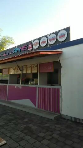 A stationary, ground-level, wide-angle shot shows a food stall called "Kuliner Rans" with a white exterior and pink and grey paneling. The stall's sign advertises various menu items like "Nasi," "Mie," "Dim Sum," and "Ayam." The stall features two windows, one labeled "Cashier" and the other "Pick Up."  The foreground includes a paved area with picnic-style wooden tables. Behind the stall are two parked motorbikes and a white building with a closed garage door. Lush green trees are visible in the background under a partly cloudy sky. The scene appears to be outdoors and deserted.