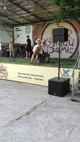 A group of people are gathered on an outdoor stage. A woman in a white shirt, yellow vest, and white cowboy hat is sitting on a chair and playing a guitar. Beside her, a man in a green uniform is standing and playing a violin. To the left, another person in a black shirt is standing. Behind them, a large banner with text and images is displayed on the stage. Two large speakers are positioned on stands on either side of the stage. The area appears to be a festival or performance venue, with a canopy overhead and a concrete ground.