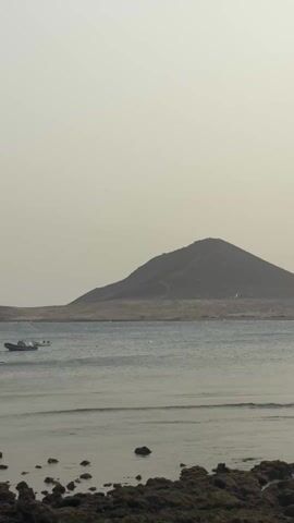 The video shows a calm ocean with a rocky shore in the foreground. In the background, there's a low mountain. There are several small boats in the water. As the video progresses, the camera pans slightly to the right, revealing a concrete pier with people standing on it. Three kites are visible in the sky above the pier, suggesting kite surfing or similar water sports activities. The sky is overcast.
