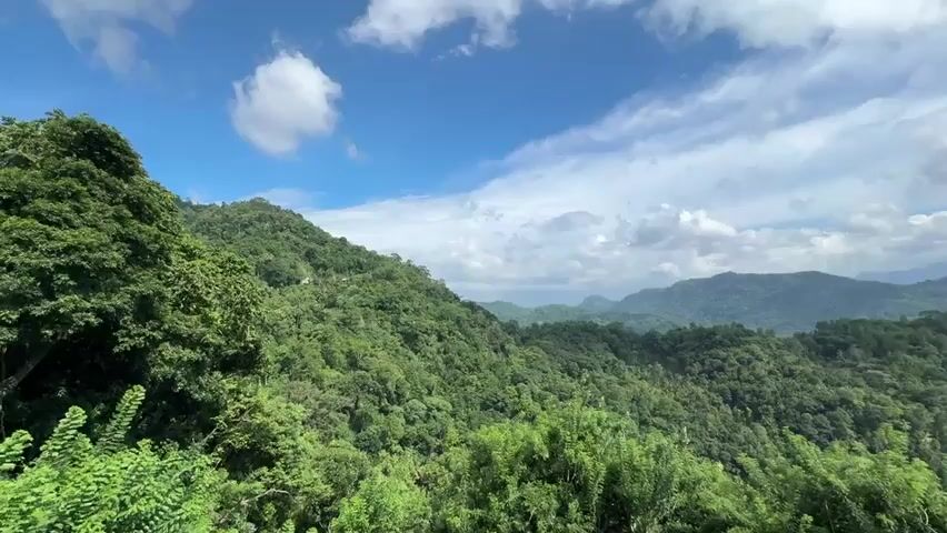 The video shows a wide view of a mountain range covered in dense green forest under a bright blue sky with fluffy white clouds. The mountains are layered and stretch into the distance. At the start of the video, there's the sound of a vehicle passing by.