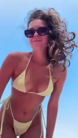 The video shows a woman wearing a light yellow string bikini and sunglasses standing outside against a bright blue sky. She initially poses, then puts her hands behind her head, and then reaches up. The camera angle is low, looking up at the woman.
