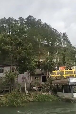 The video shows a wide river flowing through a settlement. On the left side of the river, there is a steep, tree-covered hill with buildings and yellow barriers. A small open-air structure with a white roof is situated near the riverbank. Further down the river, a large concrete bridge spans across, with a smaller red-railed bridge underneath it. On the right side of the river, there is a paved walkway or embankment. A person wearing a hooded jacket and light-colored pants is crouched on this embankment, appearing to interact with the water. The sky is overcast, suggesting a cloudy day. The water in the river is a murky green and appears to be flowing at a moderate pace, with some ripples visible. The overall scene suggests a somewhat developed area adjacent to a natural waterway.