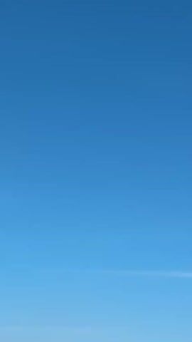 The video begins with a clear blue sky. Several white jet planes fly in formation, in the upper center of the screen, passing over a beach. Beach umbrellas and people dot the bottom of the screen, under the planes. The camera moves to follow the planes, which are the main focus of the video.