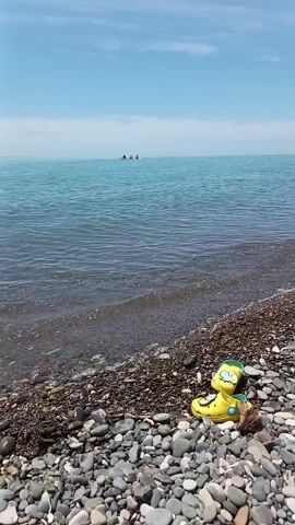 A sunny outdoor scene shows a pebbly beach with gentle waves washing ashore. The clear blue sea extends to the horizon under a light blue sky. In the mid-ground, a pair of yellow clogs with green accents rests on the dark pebbles. In the far distance on the water, several individuals are visible on what appears to be a small craft.