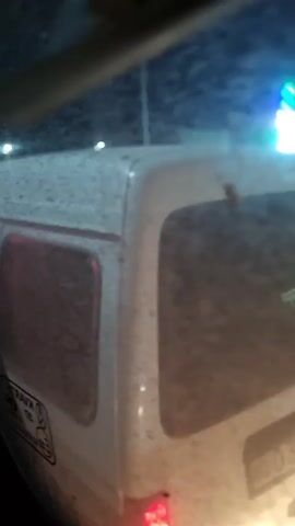 The video shows a poorly lit street scene at night, likely captured from within a moving vehicle. The main focus is on the rear of a white van, with its red taillights visible.  Other vehicles, including white cars, are also visible, along with building lights in the background, some appearing green and blue. The video quality is shaky and blurry, hindering clear detail. Some indiscernible people are seen around the white cars.