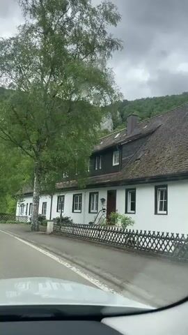 The video shows a car driving on a road in a rural area. The road is surrounded by trees and a large rock formation is visible in the distance. There is a white building with dark brown trim on the side of the road. The sky is overcast.