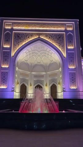The video shows a close-up view of the entrance to a building at night, with a fountain in the foreground. The building is illuminated with purple and yellow lights, highlighting its architectural details. The fountain has water spraying upwards, with colorful lights that change from pink to blue. The fountain is situated in a paved area, with steps leading up to the building's entrance. The building appears to be a mosque, with traditional Islamic architectural elements such as arches and decorative carvings. The scene is serene and majestic.