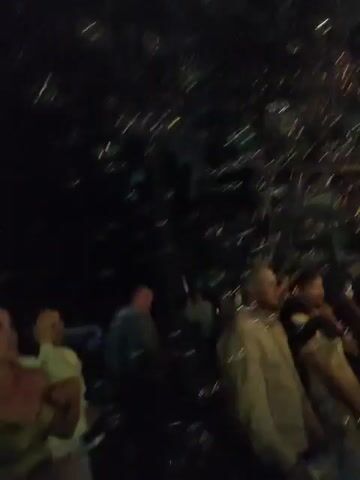 The video captures a dark outdoor scene with a blue light emitting from a device on the left, producing a stream of bubbles upwards. Several people are visible in the background, some appearing to be observing the bubbles. The lighting is dim, suggesting it is nighttime or a poorly lit area. The overall atmosphere seems festive or celebratory.