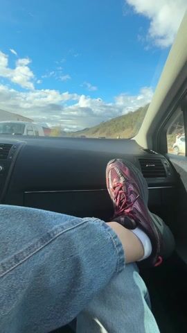 The video shows a person in the passenger seat of a car, with their leg stretched out and foot resting on the dashboard. The person is wearing blue jeans, white socks, and a burgundy and black sneaker. The background shows a sunny day with a blue sky, white clouds, mountains, and buildings. The car is moving down the road.