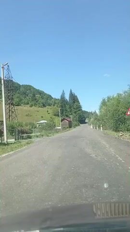 A daytime outdoor shot shows a dirt road with a car dashboard in the foreground. The road leads into a wooded area with rolling hills in the background. There are power lines to the left of the road and a small red building and some trees along the side of the road. The sky is blue with a few white clouds.