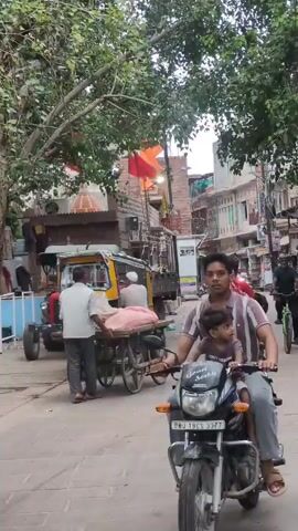 A man rides a motorcycle with a child in front of him on a street. He is wearing a striped shirt. The child is wearing a purple shirt. Behind them, people are walking and riding other motorcycles and a bicycle. A red and yellow auto rickshaw is parked on the side of the road. Trees with green leaves are overhead. Buildings line the street. A red flag with an orange symbol is visible on top of one of the buildings. The ground is paved with stone.