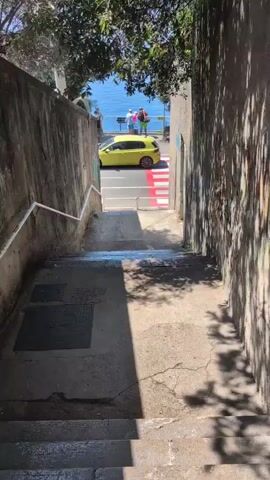 The video shows a view from the top of a set of concrete stairs looking down towards a street and the ocean. A bright yellow car is parked on the street. A red and white crosswalk is visible in front of the car. There are several people standing near the car, looking out at the ocean. The stairs are in shadow, and the street and ocean are brightly lit by the sun. The walls on either side of the stairs are made of rough stone. Trees are visible at the top of the stairs, casting shadows on the steps.