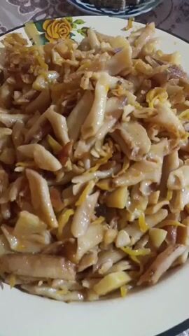 The video shows a close-up of a plate of food. The food appears to be pasta or noodles that have been mixed with vegetables and sauce. The plate is white with a floral pattern around the rim. In the background, there is a small stack of what looks like dessert, possibly halva, on a doily. A patterned tablecloth is visible beneath the plate. The lighting is dim.