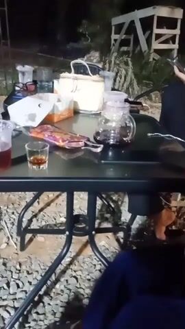 The video shows an outdoor gathering at night around a glass-topped table. A man in a green t-shirt sits on the left, holding a glass. On the table are several drinks in plastic cups, a small shot glass with amber liquid, a glass jar with a dark liquid, and a package of what appears to be tissues or wipes. A person in a dark blue shirt is visible in the lower right corner, partially obscured. The ground is covered in gravel. In the background, there is some lighting and foliage, as well as what looks like a wooden structure. The scene appears to be casual and social.