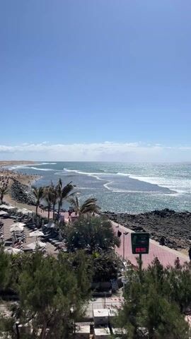 The video shows an elevated view of a sunny beach scene. The sky is clear blue with a few wispy clouds. Below, the ocean is slightly choppy with visible waves. A sandy beach stretches along the coastline with people visible enjoying the beach.  A red-tiled pathway runs along the edge of the beach, and a digital display shows the temperature as 20°C and the time as 11:39. Palm trees and other green vegetation are interspersed along the path. A rocky area borders the path and ocean.  A few people walk along the pathway.