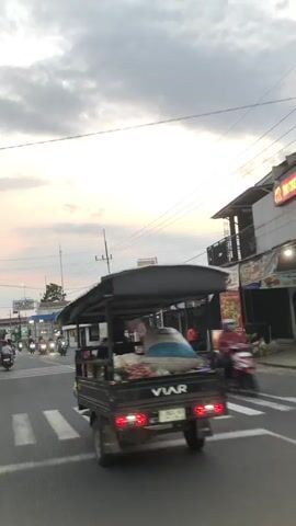 The video opens with a view of a street with a tuk-tuk driving away from the camera. The sky is cloudy with hints of pink and orange, suggesting it is either sunrise or sunset. The tuk-tuk is dark-colored and has a canopy. It appears to be carrying some goods in the back, possibly produce. There are other vehicles, including motorcycles, on the street. Buildings line the sides of the road, some of which appear to be shops or businesses. A pedestrian crossing is visible in the foreground. The overall scene suggests a moderately busy street in an urban or semi-urban environment.