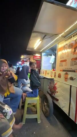 A young boy in a dark jacket and blue jeans kneels on a yellow stool in front of a food cart at night. He is looking towards the camera with a slight smile. The food cart is brightly lit with a string of white lights along the top. A menu board is visible on the right side of the cart, displaying prices in Indonesian Rupiah. Behind the boy, a woman wearing a headscarf and blue jeans is sitting, looking down at her phone. Another person is partially visible to the left. The background shows a street scene with other food stalls and people. The overall atmosphere appears to be a lively night market.