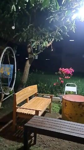 The video shows an outdoor scene at night. In the foreground, there are two wooden benches and a large, cylindrical object that appears to be a repurposed oil drum, painted yellow and red. Behind these, a wooden bench sits under a tree. Further back, a white chair is positioned near a bush with pink flowers. To the left, a white, circular swing with a blue seat is visible. The background is dark, with hints of greenery and a building structure. A bright light fixture illuminates the area from above, casting shadows. The overall atmosphere is dimly lit and appears to be a seating area for relaxation or dining.