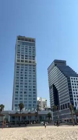 The video shows a beach with two modern buildings in the background. The first building is a tall, rectangular tower with many windows. The second building is shorter and has a unique, angular design with a large, open space in the middle. There are palm trees and streetlights along the beach. People are walking on the sand. The sky is clear and blue.