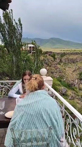 This video shows a view from a balcony overlooking a deep gorge and rolling green hills in the distance. Two people are seated at a table on the balcony, with their backs to the camera. One person, on the right, is wearing a light blue and green striped blanket around their shoulders and has blonde hair pulled back in a ponytail with a pearl accessory. The other person, on the left, has long blonde hair and is wearing a white top with light blue trim. A white table with a plate and glasses is visible between them. A large tree with green leaves and a white decorative sphere on a pillar are also on the balcony. The sky is overcast.