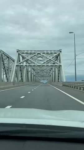 The video shows a view from inside a vehicle driving across a large, grey, metal truss bridge on a cloudy day. The road is asphalt and has white lane markings. The bridge has a repetitive geometric structure of beams and supports. In the distance, the bridge continues and the sky appears lighter. The front of the vehicle, specifically the hood and windshield, is visible in the foreground. The overall impression is one of travel over a substantial piece of infrastructure.