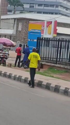 The video opens on a street scene. In the foreground, a person wearing a yellow t-shirt and black pants stands facing away from the camera, looking towards the street. The person has a medium skin tone and appears to be an adult. To their left, a concrete curb runs along the edge of the road. Behind the curb, there is a sidewalk.

On the sidewalk, a group of people are gathered. One person in a red shirt stands near the curb, facing the group. Another person in a blue robe stands in the middle of the group. There are several other individuals, some of whom appear to be seated or lying on the ground. An object resembling a shopping cart or trolley is visible near the group.

Behind the gathered individuals, a black metal fence runs horizontally across the frame. Beyond the fence, a building with signs is visible. One sign is predominantly yellow and red, with text that includes "Chicken Republic". Another sign is blue and white with text. A portion of a building with a slanted roof is visible in the upper left background. The overall environment appears to be an urban street. The lighting suggests it is daytime, possibly overcast. The video quality is grainy.