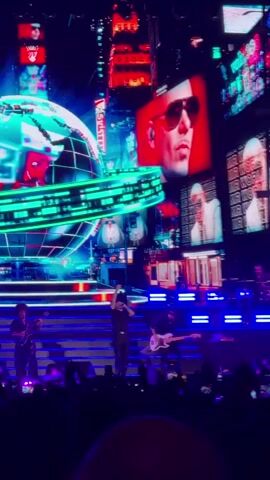 The video shows a live concert performance. In the foreground, several musicians are on stage, including a lead singer in the center wearing a black t-shirt and dark pants, and guitarists on either side. Behind them, a large, glowing, blue and green globe is prominently displayed, with a cityscape backdrop featuring illuminated buildings and screens. One large screen shows a close-up image of a man's face, wearing sunglasses, with the text "MR WORL.." visible. The stage is lit with blue and purple lights, and a crowd is visible at the bottom of the frame.