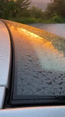 A close-up shot focuses on the wet surface of a car's hood, reflecting the warm hues of a sunset. Water droplets glisten on the dark, curved material. In the background, rolling hills are silhouetted against a vibrant orange and yellow sky. Trees frame the landscape, and a paved road is visible in the distance. The camera is positioned low, emphasizing the reflective quality of the car's surface and the expansive sky. The scene conveys a sense of tranquility and the beauty of nature at dusk.