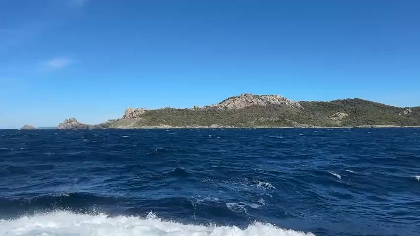 A wide shot captures a sunlit seascape with a clear blue sky overhead. The foreground is dominated by choppy, dark blue ocean water, with white foam visible where waves break. In the middle ground, a long, rocky island stretches across the frame, its landscape covered in sparse vegetation and punctuated by rugged rock formations. A prominent feature on the island's left side is a stone fortification, possibly a castle or fort, with visible walls and structures integrated into the hillside. The island's terrain slopes upwards towards the right, revealing more rocky outcrops and dense green foliage. The camera appears to be moving, likely on a boat, as evidenced by the motion of the water.