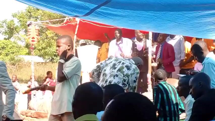 The video shows a gathering of people outdoors, possibly at a celebration or ceremony, held under a makeshift tent structure made of blue and red tarps. A group of people are gathered near the front, with one man holding a microphone and speaking. A young boy wearing a white shirt with writing on it is seen holding a drumstick and standing near the front, looking distracted or bored. There are other adults and children present, some sitting and some standing. In the background, trees and a building can be seen, suggesting the event is taking place in a rural or semi-rural location. A speaker is set up on a wooden post, presumably for the event's audio.