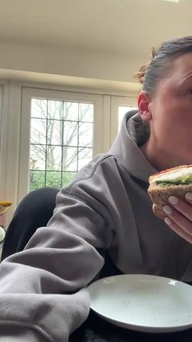 A woman with brown hair pulled back in a bun is sitting at a dark countertop in front of a white plate. She is wearing a grey hoodie and black leggings. She takes a large bite of a sandwich made with dark bread, lettuce, and other fillings. Behind her is a window showing a tree without leaves. The countertop and surrounding areas are brightly lit.