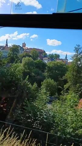 The video shows a view from a moving vehicle, possibly a train or tram, looking out a window. The foreground is dominated by lush green trees and foliage, which appear blurred due to the motion. In the background, a town or city is visible on a hill, with buildings and a steeple. The sky is blue with scattered white clouds. The overall impression is of a scenic journey through a green landscape with a glimpse of urban architecture.
