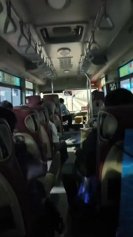 The video shows the interior of a bus from a rear-facing perspective. Several passengers are seated on the bus. The seats are upholstered in a combination of maroon and white. Overhead, there are grab handles for standing passengers. At the front of the bus, a driver is visible in the driver's seat, and some luggage or boxes are stacked near the front. The lighting inside the bus is dim, with some ambient light coming from the windows. The bus appears to be in motion, as suggested by the slight blur in the background and the movement of the interior elements.