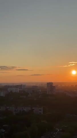 The video presents a panoramic aerial view of a city during what appears to be a sunset or sunrise. The sky dominates the upper portion of the frame, transitioning from a pale blue at the top to a vibrant orange and yellow hue closer to the horizon. The bright, circular sun is prominently visible on the right side of the frame, low in the sky, casting a warm glow. To the left, a distinct, bright, circular lens flare is also visible, mirroring the sun's position.

Below the horizon, the city stretches out, silhouetted against the bright sky. Numerous residential and possibly commercial buildings, primarily multi-story structures, are visible across the landscape. Darker patches of trees and greenery are interspersed among the buildings, especially in the foreground and mid-ground. The overall impression is that of a sprawling urban area. The architecture of the buildings suggests a location such as Dikaya Gar', Russia, as provided in the context.

The atmosphere is serene and calm, characterized by the soft, warm light of the rising or setting sun. There are a few scattered clouds near the horizon, illuminated by the sun's rays. No people or specific activities are discernible, as the view is static and from a high vantage point. There is no visible text in the video.