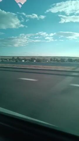 The media file depicts a view from inside a car traveling on a highway in Zhumabek, Kazakhstan. The primary subjects are the interior of the car, including seats and a dashboard, and the passing scenery outside. The scene shows a wide, paved highway with multiple lanes, bordered by a flat, arid landscape with sparse vegetation and distant structures that appear to be utility poles or possibly buildings.

The activity taking place is the journey itself, with the camera capturing the view as the car moves forward. The time of day appears to be daytime, likely late morning or early afternoon, given the bright sunlight and clear blue sky with scattered clouds. The weather seems to be fair and dry, consistent with an arid environment.

Notable details include the car's interior, which suggests a passenger's perspective. The dashboard is visible, displaying a navigation screen and radio. The sunlight is strong, casting shadows and highlighting the sky. The road is well-maintained, with clear lane markings. The distant landscape offers little in the way of distinct landmarks, reinforcing the sense of a vast, open terrain. There are no explicit emotional cues, but the imagery suggests movement and travel through a relatively unpopulated area.

No visible text is decipherable in the media file. The overall impression is one of a road trip through a somewhat desolate but scenic landscape in Kazakhstan.