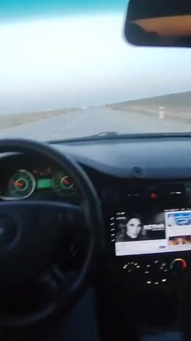 The video shows a car's dashboard and view through the front windshield and driver's side window while driving on an open road. The car's steering wheel is visible, and the driver's hand can be seen holding the wheel. A dashboard screen plays a video with a woman on it. The landscape seen through the windshield and side window appears flat and desert-like, with wind turbines visible in the distance. The sky is light, suggesting it could be dawn or dusk.