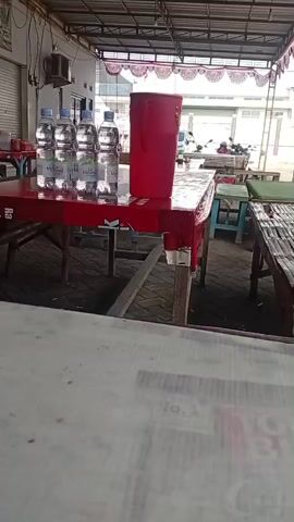 The video shows an outdoor dining area with several tables and seating. A red-covered table in the foreground has six bottles of water and a tall red cup on it. Behind this table, other tables with chairs are visible. The environment appears to be a casual eatery or roadside restaurant, with a roof providing shade and a closed metal shutter on a building in the background. A motorcycle is parked to the right. The camera is positioned at a low angle, looking across a white surface, likely another table, at the scene.