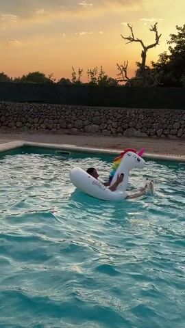A person is in a swimming pool on a large inflatable unicorn. The person is partially submerged in the water and appears to be holding onto the unicorn's neck. Water is splashing around the person and the unicorn. The sun is setting in the background, casting an orange glow over the scene. There are trees and a stone wall visible behind the pool.
