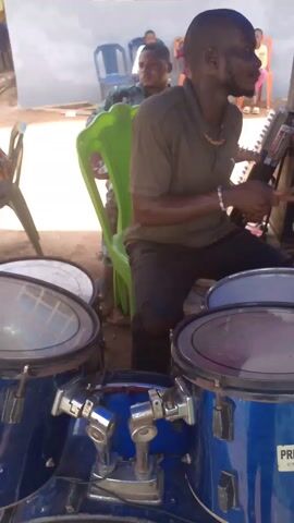Here's a description of the video:

The video shows a man playing a drum set outside, likely at an event or gathering. He is seated on a green plastic chair, wearing a brown t-shirt, dark pants with a rip in the knee, and a beaded necklace. He's focused on playing the drums, using drumsticks to hit the cymbals and drums. The drum set is blue and appears to be a Premier brand.

Behind the drummer, there are speakers stacked on top of each other and a keyboard. Other people are seated in chairs watching or listening to the music. The setting appears to be outdoors, possibly a yard or event space near a building with white walls and visible windows.